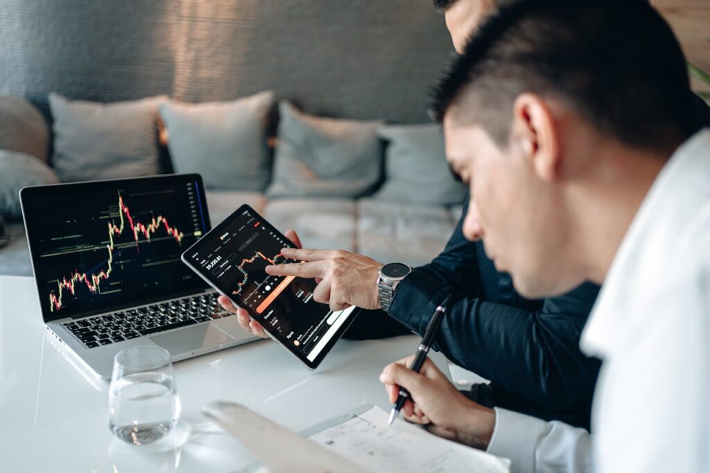 Two businessmen analyzing stock charts on a laptop and tablet in an office setting.